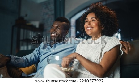 Couple Watching Comedy Movie on TV, Eating Popcorn while Sitting on Couch in the Apartment Late at Night. Laughing Boyfriend and Girlfriend Enjoying Funny TV Series at Home. Low Angle Shot.