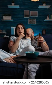 Couple Watching Scary Horror Movie On Television Channel, Feeling Afraid And Terrified In Living Room. Woman With Mouth Open And Man Hiding Behind He, Looking At Film On TV Program. POV