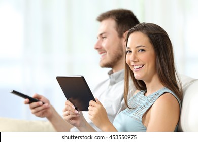 Couple Watching Tv Sitting On A Sofa In The Living Room At Home. Man Is Zapping And The Woman Is Holding A Tablet And Looking At You