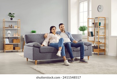 Couple watching TV together while sitting on couch in living room. Cheerful spouses sitting hugging on comfy sofa watching television. Happy family spending time together at home - Powered by Shutterstock