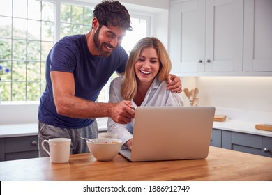 Couple Wearing Pyjamas In Kitchen Watching Laptop Whilst Eating Breakfast