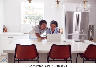 Couple Wearing Pyjamas Standing In Kitchen Working From Home On Laptop