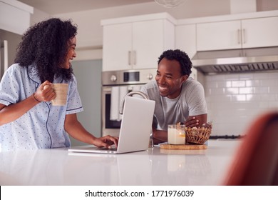 Couple Wearing Pyjamas Standing In Kitchen Working From Home On Laptop