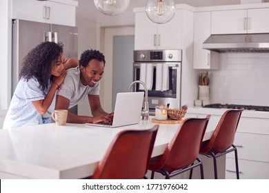 Couple Wearing Pyjamas Standing In Kitchen Working From Home On Laptop