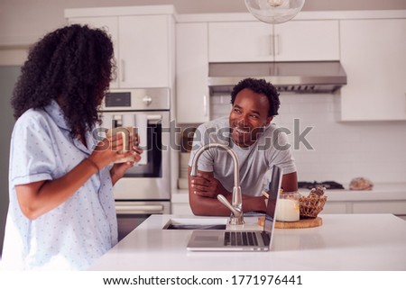 Couple Wearing Pyjamas Standing In Kitchen Working From Home On Laptop