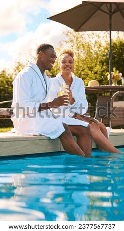 Couple Wearing Robes Outdoors Sitting With Drinks With Feet In Swimming Pool On Spa Day