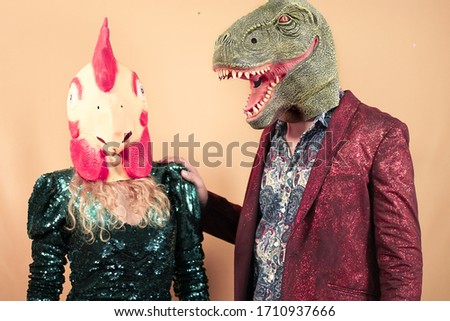 Couple wearing with t-rex dinosaur and chicken mask. Crazy friends having fun celebrating a party. Absurd and surreal funny concept. Yellow background - Image
