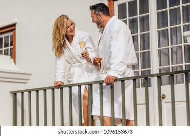 Couple wearing white bath robes standing on balcony and smiling at each other. Smiling man and woman in bathrobes having wine and talking.