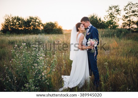 couple in wedding attire with a bouquet of flowers and greenery is in the hands against the backdrop of the field at sunset, the bride and groom