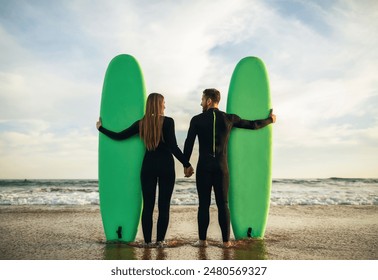 A couple in wetsuits are standing on a sandy beach, holding green surfboards. They are facing each other, holding hands. The ocean is behind them, and the sun is setting in the background. - Powered by Shutterstock
