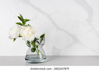 A Couple Of White Peony Flowers Being Placed In A Transparent Glass Vase Against A Marble-veining Kitchen Backsplash Which Was Made Of Quartz Stones