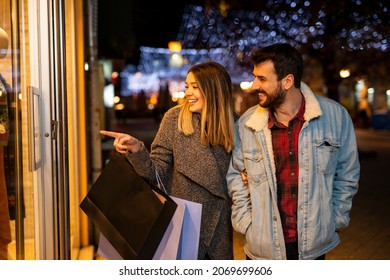 Couple window shopping outdoors in winter city street. Standing in front of a store window and looking inside. - Shutterstock ID 2069699606
