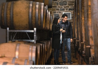 Couple In The Wine Cellar, Enjoying Drinking White Wine While Looking At Old Wooden Barrels For Wine Storage And Aging