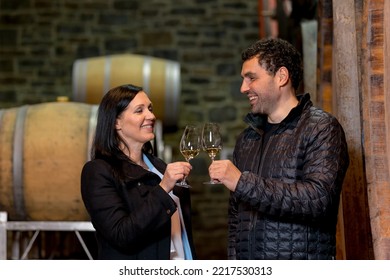 Couple In The Wine Cellar, Enjoying Drinking White Wine While Looking At Old Wooden Barrels For Wine Storage And Aging