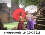 Couple woman, Asian and Caucasian friend waring Thai traditional dress travel in Ayutthaya historical park, a world heritage site. Happy travel with friend, Happy smiles, Thai hand made umbrellas