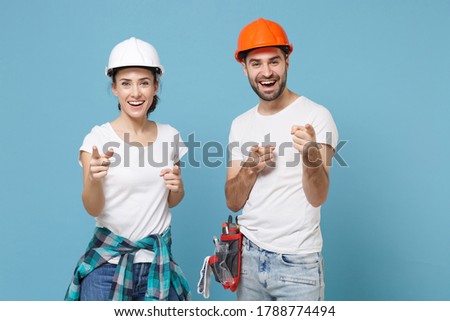 Couple woman man in casual clothes protective helmet hardhat isolated on blue background. Instruments accessories for renovation apartment room. Repair home concept. Pointing index fingers on camera
