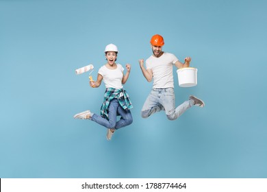 Couple woman man in protective helmet hardhat jump with paint bucket, roller isolated on blue background. Instruments accessories renovation apartment room. Repair home concept. Doing winner gesture