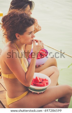 couple  women friends  sit on wooden dock by river or lake enjoy eating slices of watermelon hot summer day