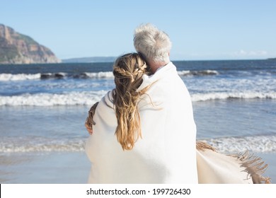 Couple wrapped up in blanket on the beach looking out to sea on a sunny day - Powered by Shutterstock