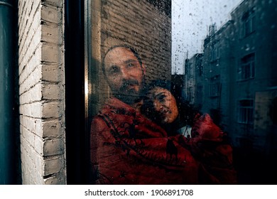 Couple Wrapped In A Red Blanket Looking Outside From A Backyard Door Window On A Grey Rainy Day.