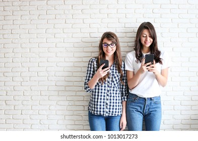 Couple Of Young Attractive Women Chatting Messaging On New Cell Phone. Two Pretty Beautiful Holding Mobile Gadget In Hands, Having Fun, Laughing & Smiling. Brick Wall Background, Copy Space, Close Up.