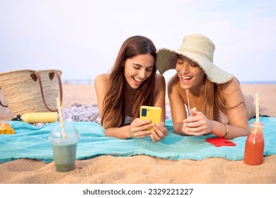 Couple of young beautiful girls looking at smiling mobile phone lying on beach. Two happy women friends use cell applications on summer holiday. Generation z cheerful people and social networks. - Powered by Shutterstock