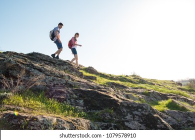 Couple Of Young Happy Travelers Hiking With Backpacks On The Beautiful Rocky Trail At Warm Sunny Evening. Family Travel And Adventure Concept.