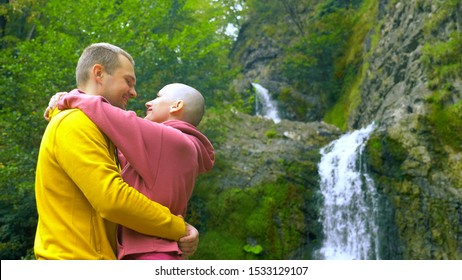 Couple, A Young Man And A Bald Girl Hug And Kiss On The Background Of A Waterfall. Travel And Family Vacation Concept. Copy Space