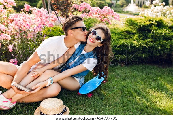 夏の公園の花の背景に草を楽しむ若者が2人いる 白いtシャツを着たハンサムな男が 長い巻き毛のきれいな女の子にキスをする 彼女は生き生きと笑う の写真素材 今すぐ編集