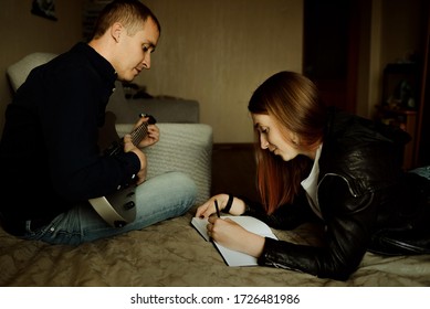 A Couple Of Young People Play Guitar At Home. A Guy And A Girl Write Music At Home. A Couple In Love On The Bed Are Engaged In Writing A Song.