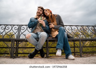 Couple Of Young People Sharing Headphones Listening To Music Together Sitting On A Bench In The Park