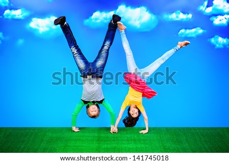 Couple of young people are standing on their heads on a green lawn over blue sky.