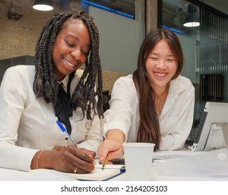 Couple Of Young Smiling Girls In An Office. African American Girl Writing In A Diary And Asian Girl Pointing At Her. Bussiness Meeting.