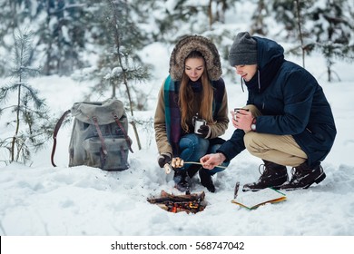 Couple Of Young Travelers Roasting Marshmallows Over Bonfire In Snowy Winter Forest