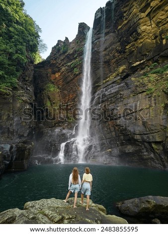A couple of young women stand against a tall tropical waterfall and hold hands enjoying the view