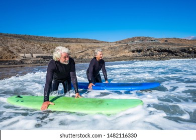 Couple Of Youth Active Seniors At The Beach With Black Wetsuits Holding A Surftable Ready To Go Surfing  - Active Mature And Retired People Doing Happy Activity Together In Their Vacations Or Freetime