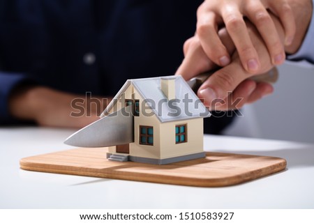 Couple's Hand Cutting The Miniature House Model With Knife On Chopping Board