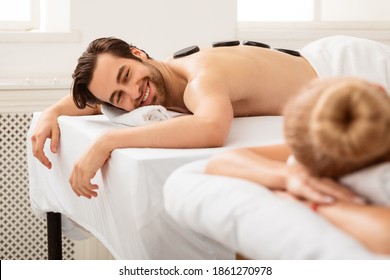 Couples Hot Stone Massage. Contented Husband Smiling To Wife Lying And Relaxing On Massage Therapy At Spa Salon During Romantic Honeymoon. Beauty, Relaxation And Wellness. Selective Focus