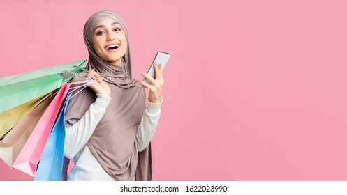 Coupon And Discount App. Happy Arabic Woman In Hijab Holding Smartphone And Paper Shopping Bags, Enjoying New Application, Panorama