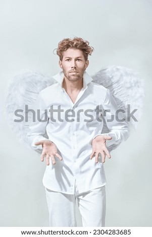 Courageous handsome male Cupid angel in white clothes and with white wings. White background. God of love Cupid. Valentine's Day.