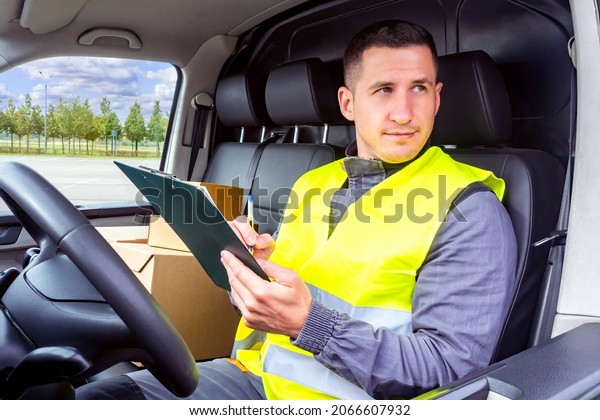 Courier in car. Delivery man in driver\'s seat.\
Male courier delivers boxes. Clipboard in hands of delivery man.\
Postman with boxes. He is engaged in delivery of parcels. Working\
in postal service