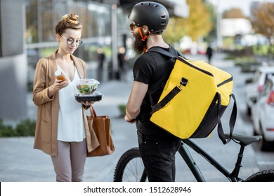 Courier delivering fresh lunches to a young business woman on a bicycle with thermal backpack. Takeaway restaurant food delivery concept - Shutterstock ID 1512628946