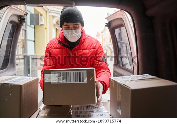 Courier delivers parcels to clients on\
car. Vertical view of the delivery man in protective mask unloads\
out of car parcel, near entrance of house. Stock\
photo