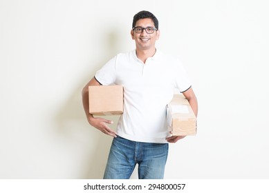 Courier Delivery Service Concept. Happy Indian Man Received Brown Boxes, Standing On Plain Background With Shadow. Asian Handsome Guy Model. 