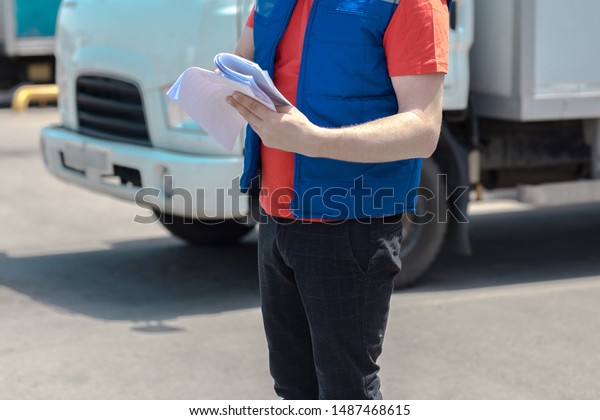 Courier driver in uniform making notes in
document and delivery white truck behinde
him