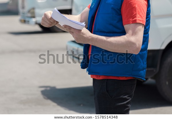 Courier driver in uniform making notes in\
document and delivery white truck behinde\
him