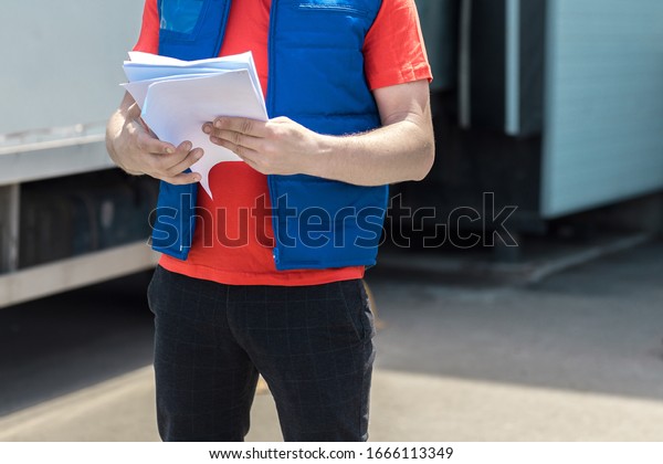 Courier driver in uniform making notes in\
document and delivery white truck behinde\
him