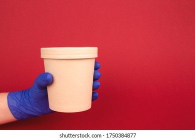 Courier Hand In Blue Disposable Protective Gloves On Burgundy Red Background, Holding Round Paper Food Container - Cardboard Cup For Soup, Ice Cream Or Other Dishes To-go. Copy Space For Text. Mockup