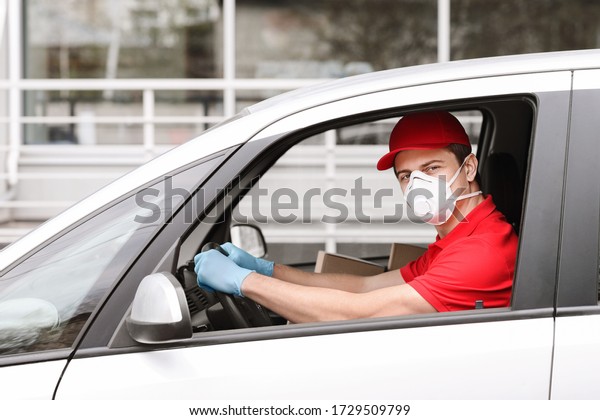Courier job\
during quarantine concept. Delivery man in red uniform in medical\
mask and gloves driving car,\
outdoors