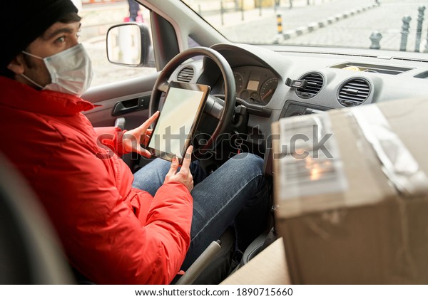 Courier job during quarantine concept.\
Delivery man in red uniform wearing medical mask looking at the\
window while driving car, outdoors. Stock\
photo
